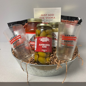 A metal basket holing two glasses bearing the GCM logo, jars of olives and pickles, bloody Mary mix, and two packs of snack sticks
