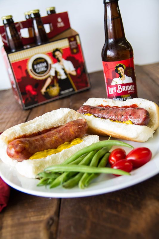 Two brats in buns with ketchup and mustard on a plate with green beans and tomatoes. Bottles of Lift Bridge beer are sitting in the background