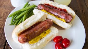Two brats in buns with ketchup and mustard sitting on a white plate along with green beans and cherry tomatoes